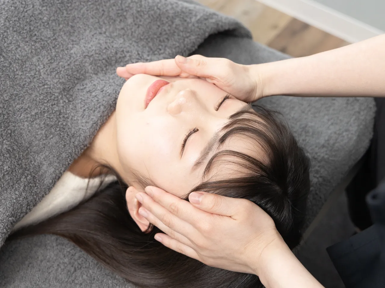 女性の頬に手を添えて小顔矯正を行う施術風景。表情筋の緊張を和らげ、フェイスラインを整える様子。